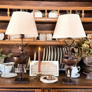 Matching pair of deer-themed bronze table lamps with beige shades displayed on wooden hutch with candles and holiday accents