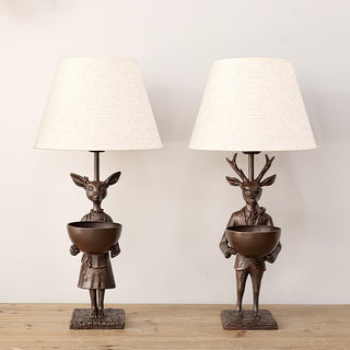 Set of two bronze deer lamps with beige shades on wooden tabletop, featuring male and female reindeer figures in elegant attire