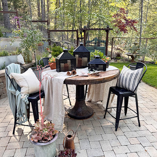 Outdoor patio dining setup featuring black iron lanterns as centerpiece décor on a round wooden table surrounded by garden greenery.