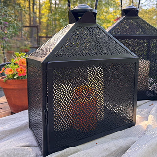 Close-up of black perforated metal lantern with pillar candle inside, styled on a table with potted flowers in an outdoor garden setting.