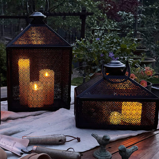 Black iron lanterns illuminated with warm LED candles at dusk, displayed on a rustic outdoor table with gardening tools.