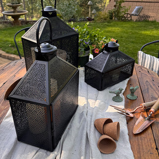 Three decorative black metal lanterns arranged on an outdoor wooden table with gardening supplies and patio greenery.