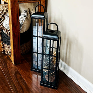Pair of black glass decorative lanterns filled with pinecones displayed beside a wooden console table on hardwood flooring