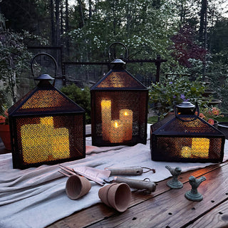 Set of three black perforated metal lanterns glowing with LED candles during twilight on an outdoor garden table.