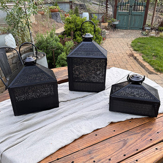 Three black metal lanterns displayed outdoors on a wooden table, showing size comparison and detailed perforated pattern.