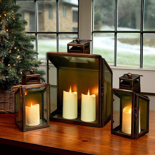Set of bronze metal and glass decorative lanterns with pillar candles displayed on a wooden table beside a lit Christmas tree, styled for cozy holiday home décor