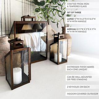 Wall-mounted bronze lanterns with pillar candles styled on a plaid-pattern backdrop alongside rustic vases and greenery for elegant home décor.