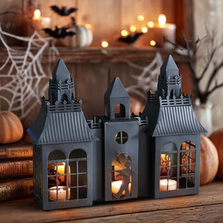 Black haunted house metal candle holder with tealight candles glowing inside, Halloween decor with pumpkins, cobwebs, and bats in background