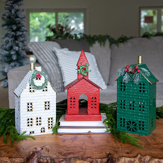 Set of three decorative Christmas metal houses — white, red, and green — displayed on a wooden coffee table with holiday greenery