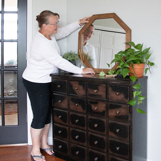 Person positioning an octagon wood-framed mirror on top of a vintage dresser with plants and reed diffuser
