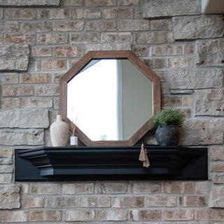 Octagon wood-framed mirror styled on a black mantel shelf against a stone accent wall with decorative vases and greenery.