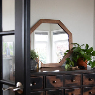 Octagon wooden mirror placed on a distressed dresser near a glass door, surrounded by greenery and home décor accents.