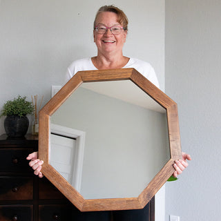 Person holding an octagon wood-framed mirror in a bright room, showing size and scale for product display.