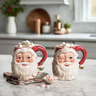 Two Santa Claus face mugs with winking expressions on marble kitchen counter with Christmas decor and hot chocolate marshmallows