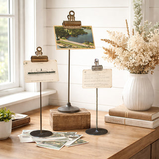 Three vintage metal note clip holders displaying photos and postcards on farmhouse table with books and dried flowers