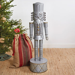 Full view of 4 foot tall galvanized metal nutcracker soldier with classic uniform drum base and weathered silver gray finish next to wrapped Christmas presents