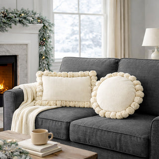 Cream pom pom decorative pillows displayed on gray sofa in cozy living room with fireplace and holiday decor