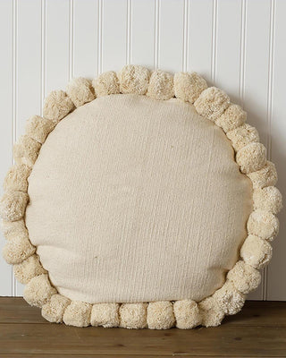 Round cushion with pom-pom trim on a wooden surface against a white paneled wall.