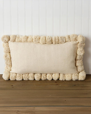 Beige pillow with pom-pom trim on a wooden surface against a white paneled wall.