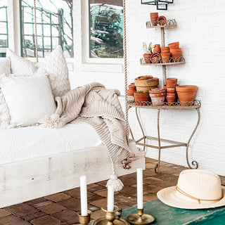 Cozy outdoor seating area with a white cushioned bench, a plant stand, potted plants, and a hat on a table.
