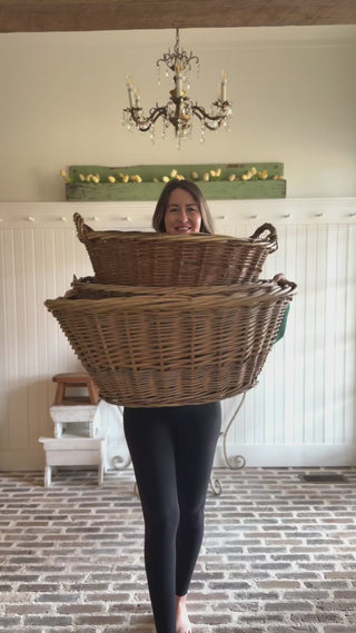 A woman styling an entryway with a beadboard accent wall with rustic decor elements including a wooden green shelf with white tulips, a plant stand with terracotta pots, a stack of vintage stools with a vintage copper pitcher, a peg rail with a hanging wicker basket and sunhats, and a chandelier.