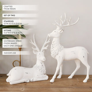 Set of two white distressed resin deer figurines, one standing and one sitting, displayed on wooden tabletop with candle and greenery