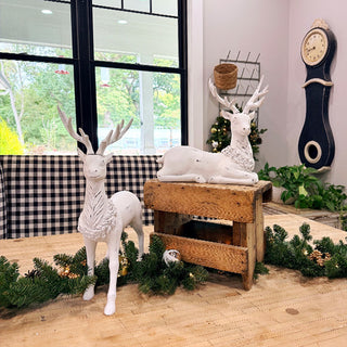 Rustic farmhouse holiday decor featuring two white resin reindeer statues on wooden table with greenery garland and vintage clock in background