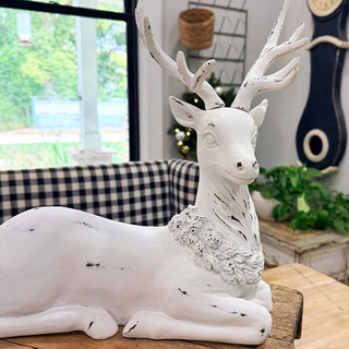 Sitting white resin deer statue with distressed finish and wreath collar, displayed on wooden table in farmhouse holiday setting