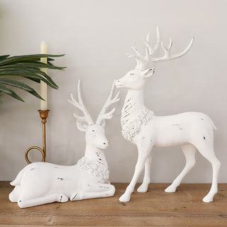 Pair of white distressed resin deer figurines—one standing, one sitting—styled on wood surface with brass candleholder and green foliage