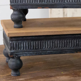 Close-up detail of wood riser showing natural wood top, black metal corrugated base, and ball-shaped feet with distressed finish