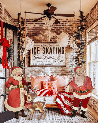 A cozy holiday-themed room decorated with Santa Claus and reindeer figures, a red-and-white striped throw blanket, a brick wall, and a large vintage-style 'Ice Skating Skate Rentals' sign above a bench.