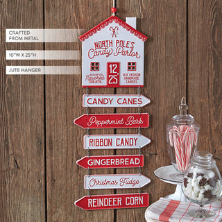 Vintage Christmas metal sign showing North Pole Candy Parlor house with directional arrows for candy canes peppermint bark gingerbread on rustic wood background