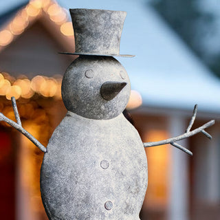 Farmhouse metal snowman decoration with top hat and carrot nose against warm holiday lights background