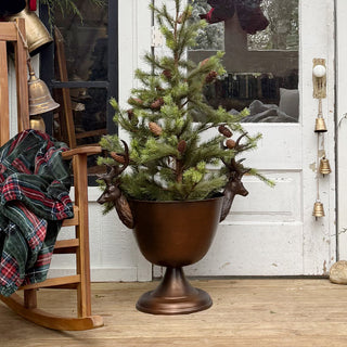 Holiday-decorated cabin porch featuring bronze stag pedestal bowl styled with evergreen tree and rustic Christmas bells.