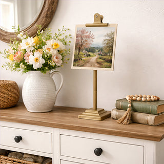 Gold metal photo stand with clip holder displaying landscape print on white dresser with flowers
