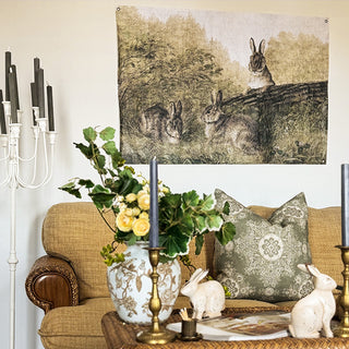 Vintage rabbit tapestry displayed above sofa with candelabra and green throw pillows