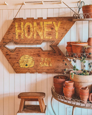 A plant stand with terrcotta pots and a honey sign including a stack of vintage stools and a pegrail in front of a beadboard accent wall