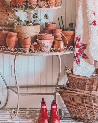 A plant stand with terrcotta pots including a vintage quilt next to wicker baskets and red rainboots in front of a beadboard accent wall