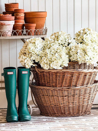 A plant stand with terracotta pots next to wicker baskets with white hydrangea s and green rainboots in front of a beadboard accent wall