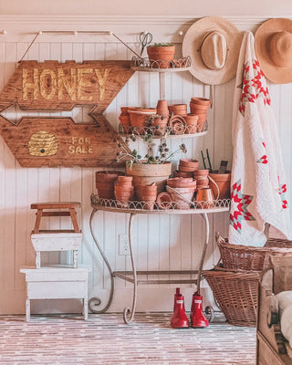 A plant stand with terrcotta pots and a honey sign including a stack of vintage stools a pegrail with a hanging sunhats and a vintage quilt next to wicker baskets and red rainboots in front of a beadboard accent wall