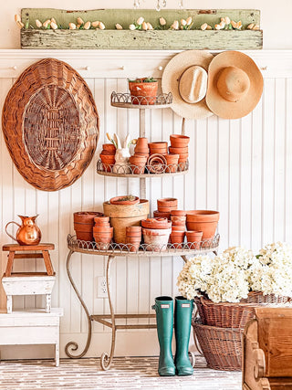 A plant stand with terrcotta pots and a bunny figurine on it including a wooden shelf with white tulips a stack of vintage stools with a vintage copper pitcher a pegrail with a hanging wicker basket and sunhats next to wicker baskets with white hydrangeas and green rainboots in front of a beadboard accent wall