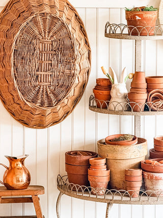 A plant stand with terracotta pots and a bunny figurine on it next to a hanging wicker basket and a vintage copper pitcher in front of a beadboard accent wall