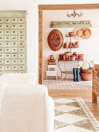 Nestled corner of a room with rustic decor elements including a wooden shelf, a plant stand with pots, a vintage cabinet, and a chandelier in front of a beadboard accent wall.