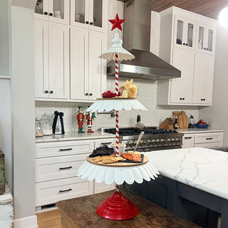 Large tiered serving stand with snacks and cheese displayed on kitchen island with white cabinets and stainless-steel range hood