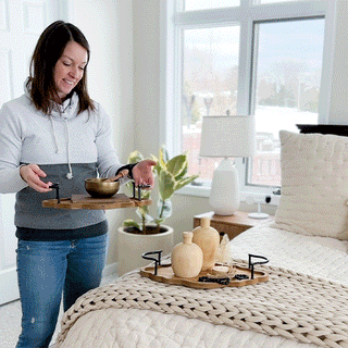 Woman styling scalloped edge wood and iron decorative tray on bedroom bed with candles and decor