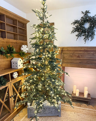 A decorated Christmas tree with warm white string lights stands in a rustic indoor room with wooden cabinetry, a wreath on the wall, and candles on a small table.