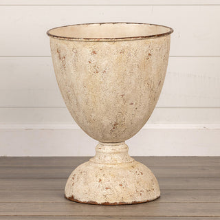 Full view of distressed cream metal urn planter on pedestal base against white shiplap wall showing classic goblet silhouette