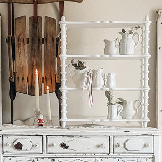 White spindle-style wooden shelf displayed on a vintage dresser with white ceramic pitchers and candles in a rustic farmhouse room.