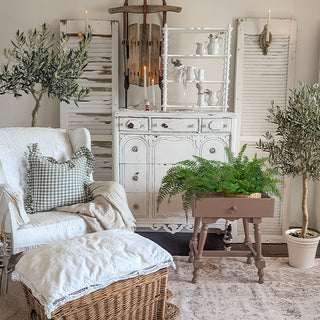 Cozy farmhouse living room featuring a white distressed dresser, wooden tiered shelf, vintage shutters, greenery, and cottage-style seating.